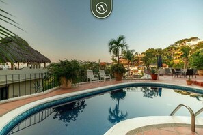 Une piscine avec des chaises longues, une structure à toit de chaume et une vue sur les bâtiments en arrière-plan.