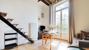 Un salon moderne avec un escalier en bois, un coin repas avec une table et des chaises, un canapé et une grande fenêtre donnant sur des immeubles.