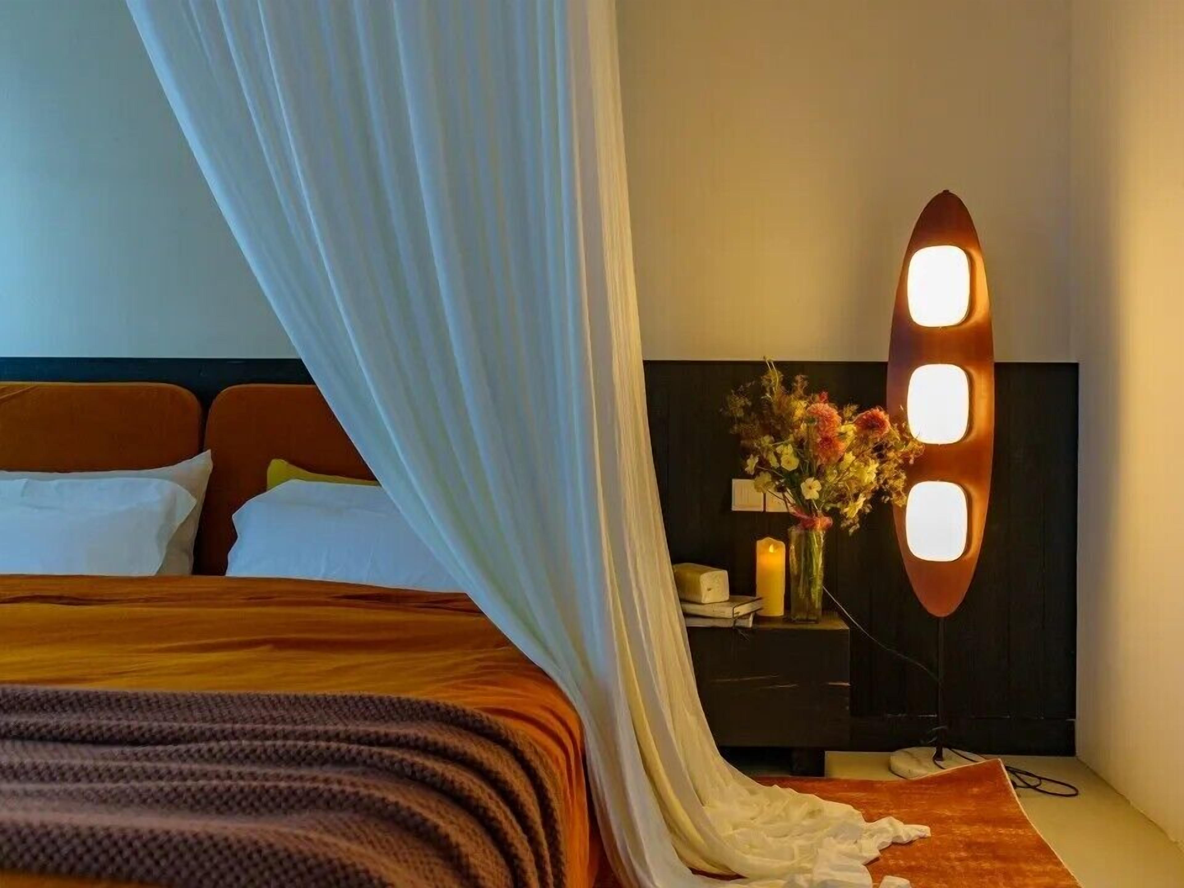 A hotel room with a bed, a bedside table with a vase of flowers, and a wall-mounted light fixture.