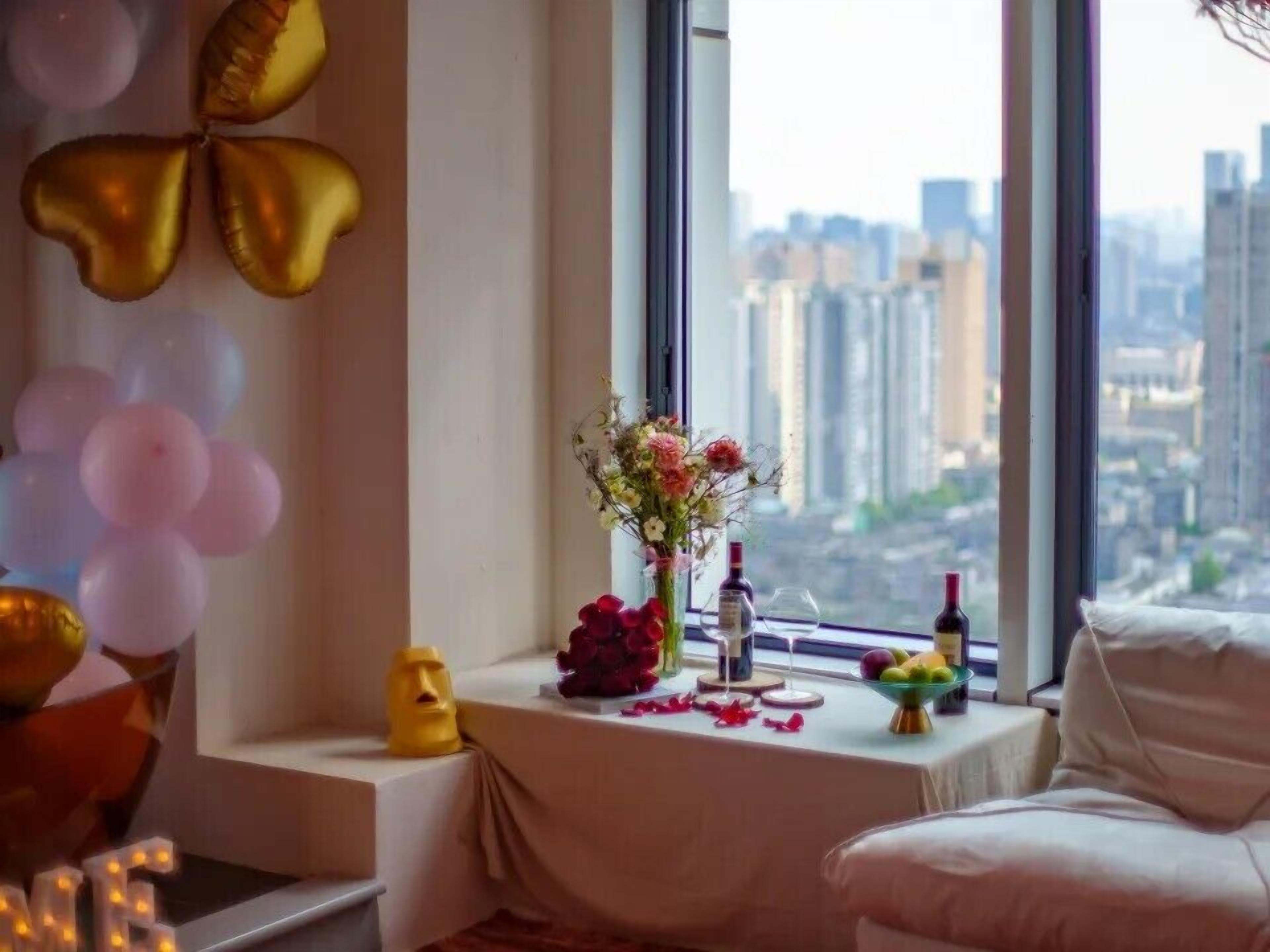 A room with a view of a cityscape through large windows, a small table with a vase of flowers, and balloons on the left side.