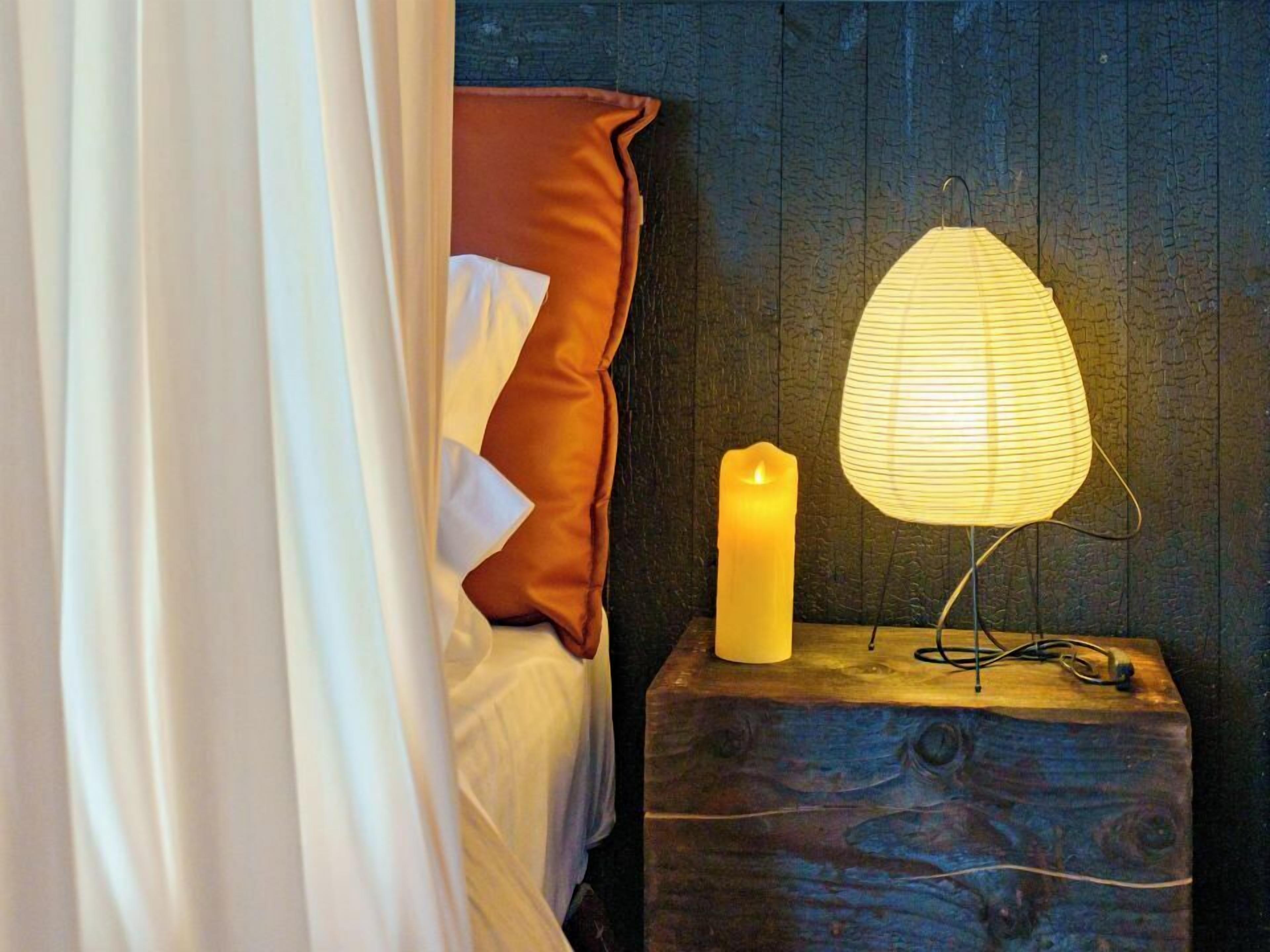 A bed with a white and brown pillow, a lit candle, and a paper lantern on a wooden nightstand.