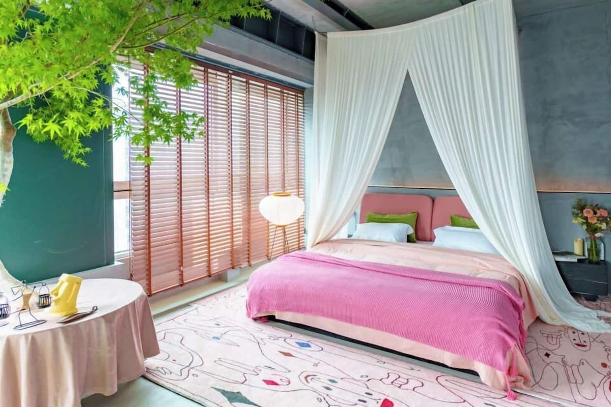 A bedroom with a bed, a bedside table, a lamp, and a window with blinds.