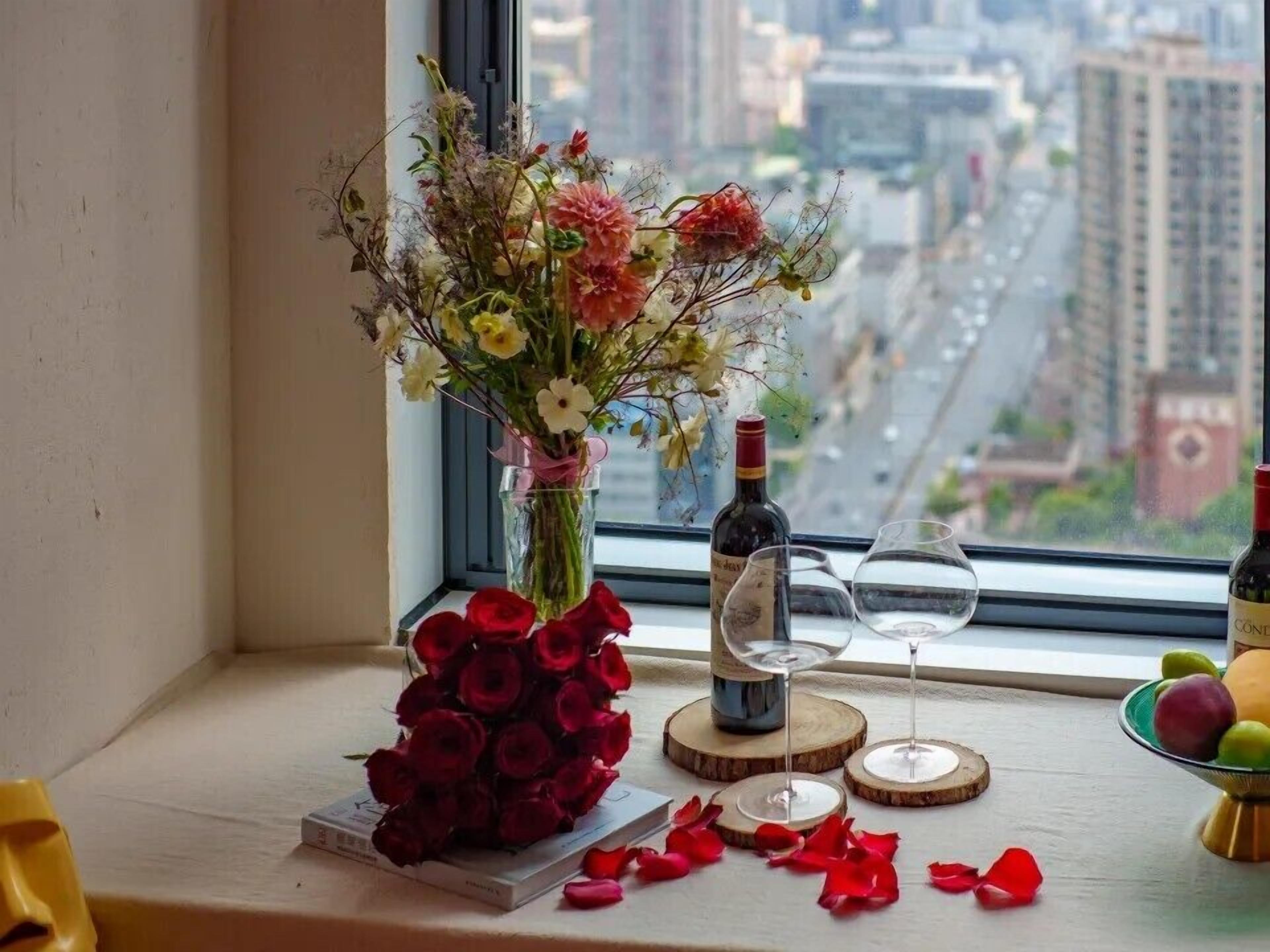 A window with a view of a cityscape, a vase with flowers, a bottle of wine, and glasses on a windowsill.