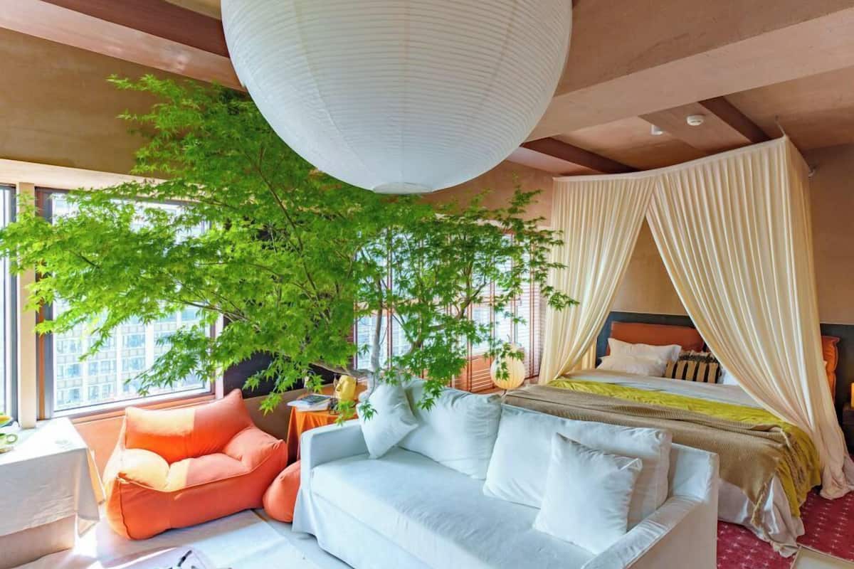 A modern bedroom with a large tree, a white sofa, a bed with a canopy, and a view of the city.