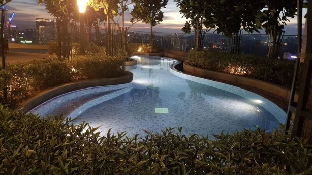 A modern infinity pool with a curved edge, surrounded by lush greenery and a cityscape view at sunset. - The Robertson Residences Free Gym & Pool (Kuala Lumpur)