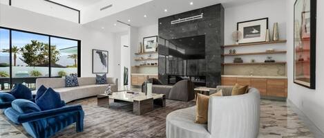 A spacious living room with a large window, a fireplace, and a kitchen area with marble countertops.
