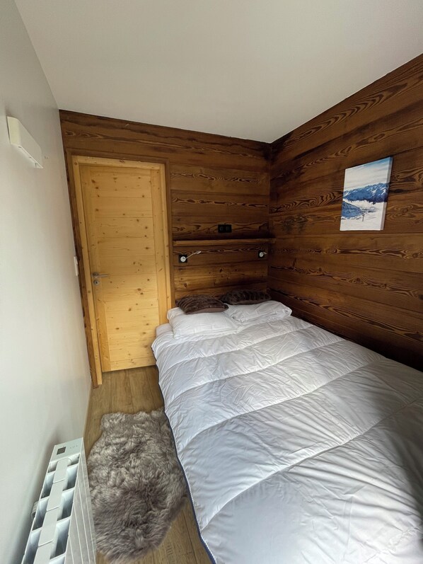 Una habitación pequeña y acogedora con paredes de madera, una cama con ropa blanca, un tapete mullido y un cuadro de una montaña nevada en la pared.