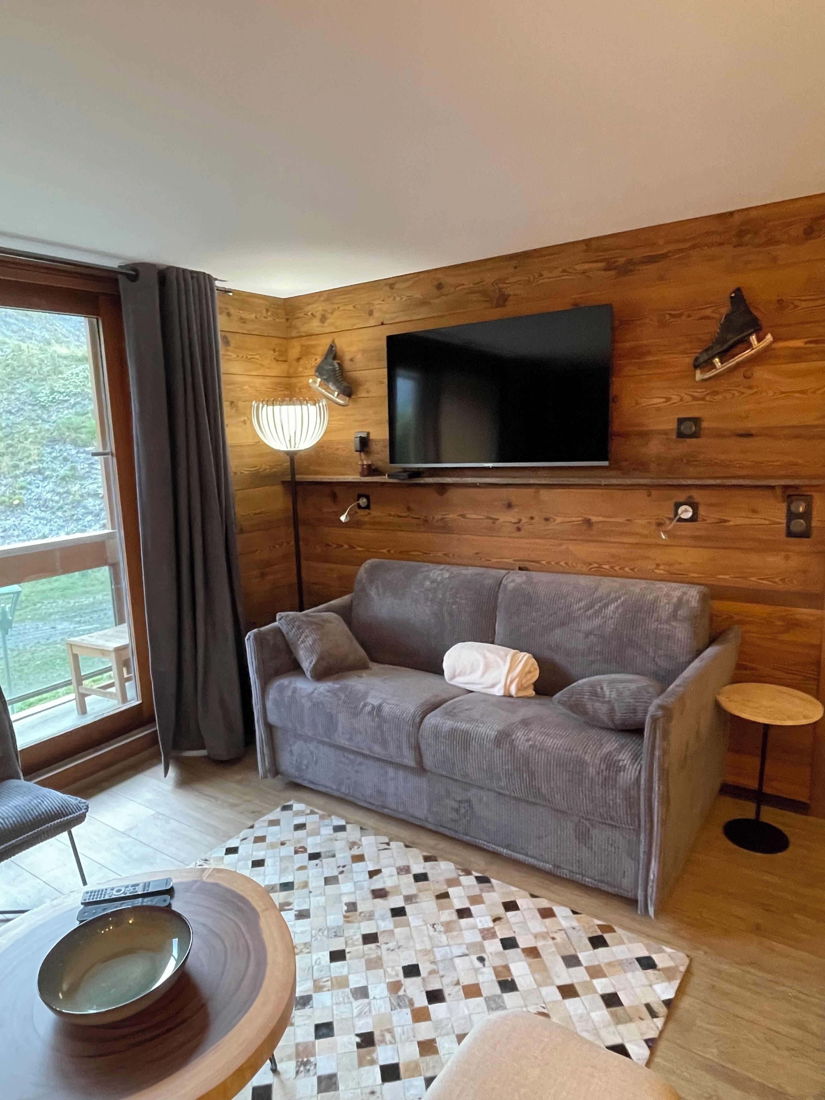 A cozy living room with a grey sofa, a flat-screen TV mounted on a wooden wall, a small round table with a bowl, and a checkered rug.