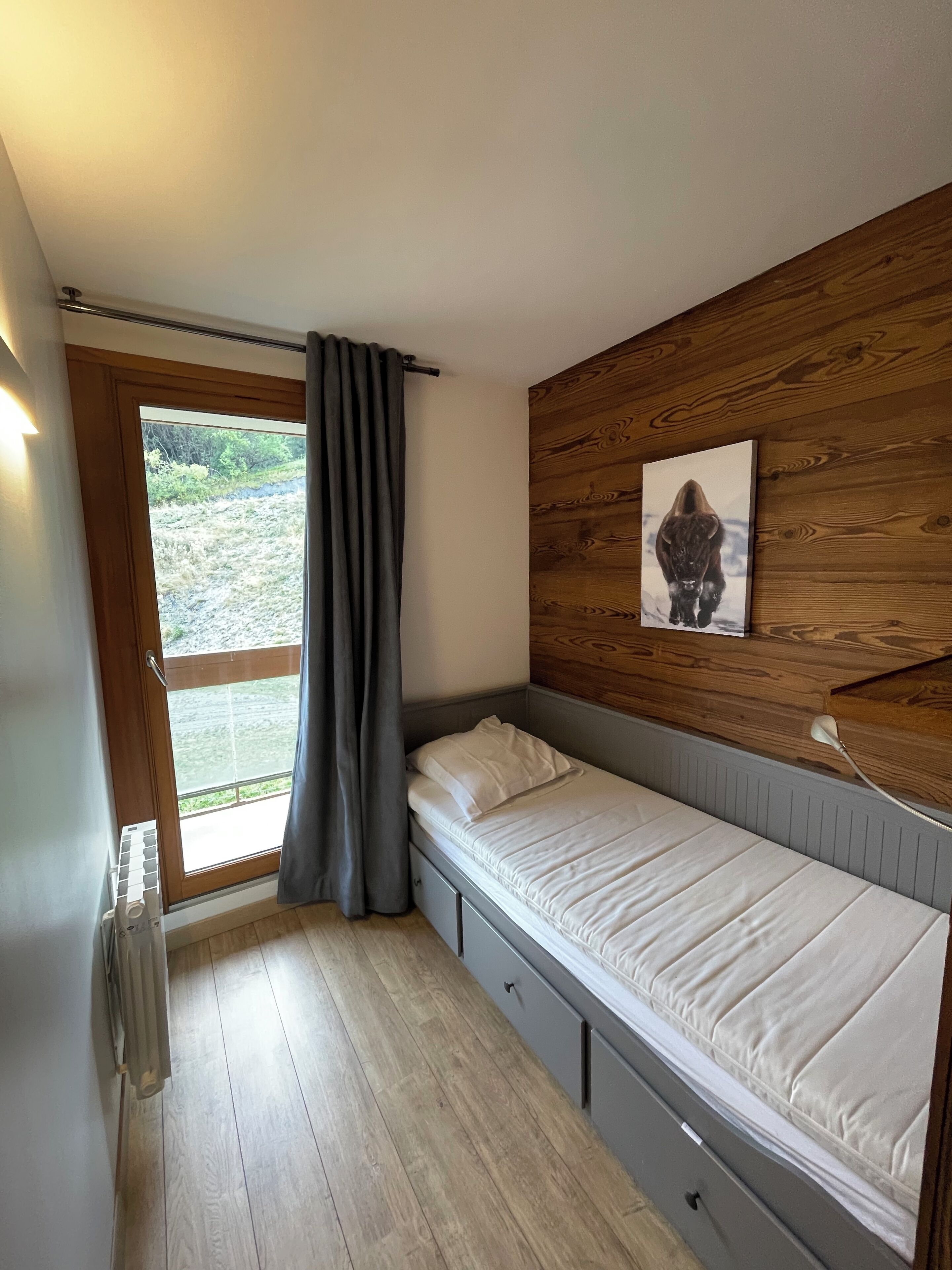 Een slaapkamer met een houten hoofdbord, een bed met witte beddengoed, een radiator, een raam met gordijnen, en een schilderij aan de muur.