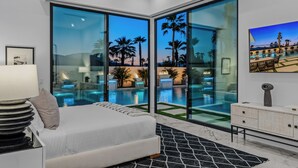 A modern bedroom with a large bed, a nightstand, a lamp, and a view of a swimming pool with palm trees.