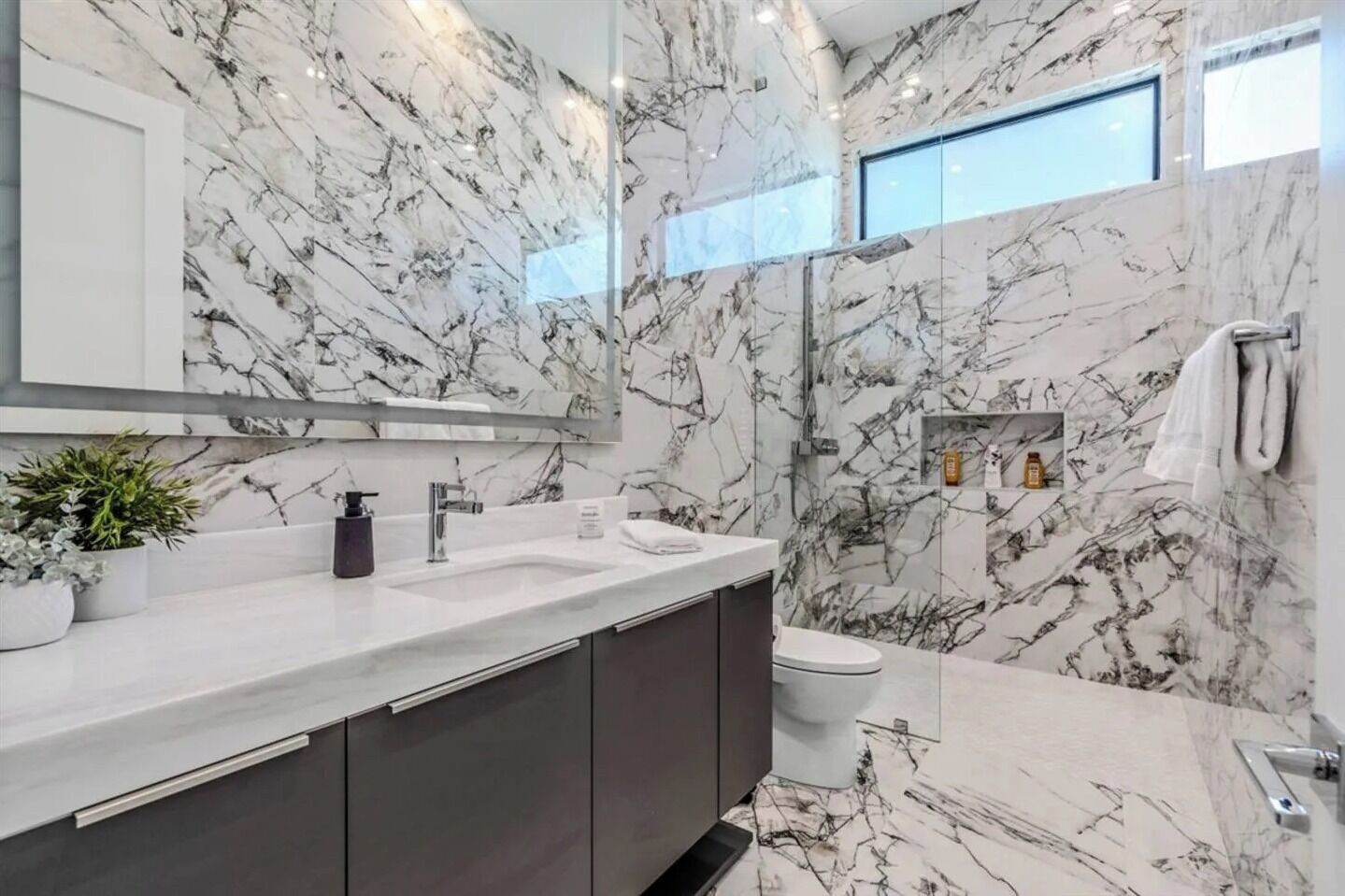 A modern bathroom with marble walls, a large mirror, a sink, and a toilet.