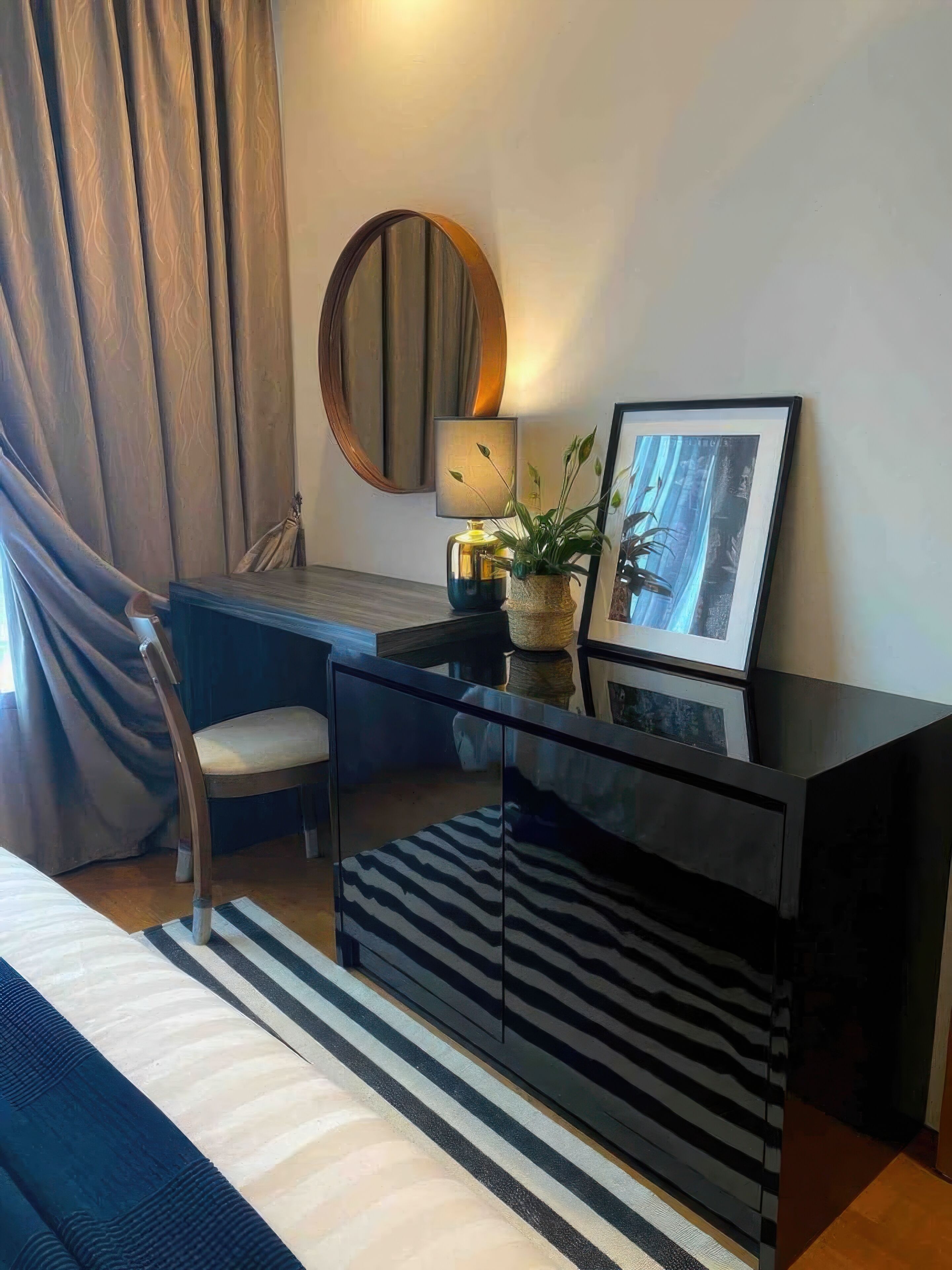 Ein modernes Hotelzimmer mit Glastisch, Stuhl, Bett, gestreiftem Teppich, runder Spiegel, Lampe, gerahmtem Bild und Topfpflanze.