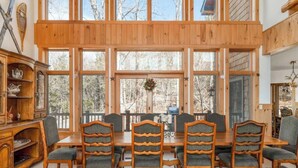 A wooden dining room with a large table and chairs, large windows, and a view of trees. - Luxe 8BR Windham Lodge | Theater, Walk to Slopes (Windham)