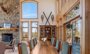 A spacious dining area with a long wooden table, stone fireplace, and large windows. - Luxe 8BR Windham Lodge | Theater, Walk to Slopes (Windham)