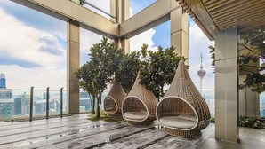 Een modern interieur met een hoge plafond, grote ramen en uitzicht over de stad. Drie rieten stoelen met kussens.