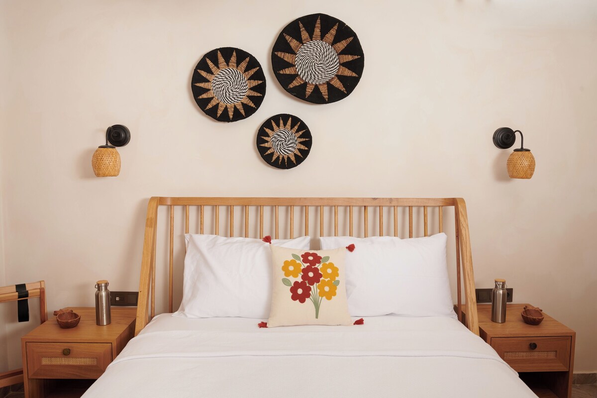 Ett välordnat hotellrum med en säng i trä, vita sängkläder och en prydnadskudde med blommotiv. Ovanför sängen hänger tre dekorativa föremål på väggen, och det finns två små sidobord med lampor och en flaska på varje.
