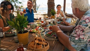 Un grupo de personas cenando al aire libre en una mesa de madera, con varios platos, una planta en maceta y una cesta de pan.