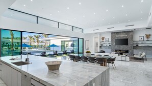 A modern kitchen with a large island, a dining area with chairs, a TV mounted on the wall, and a view of an outdoor pool area.