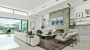 A modern living room with a fireplace, a large TV, and a view of a pool and greenery outside.