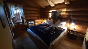 Una habitación tipo cabaña de madera con una cama, una mesita, una mesilla de noche, una ventana con cortinas y un cuadro en la pared.