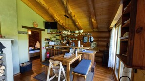 Interior rústico de una cabaña con una mesa de madera, banco y una zona de cocina con utensilios colgando.