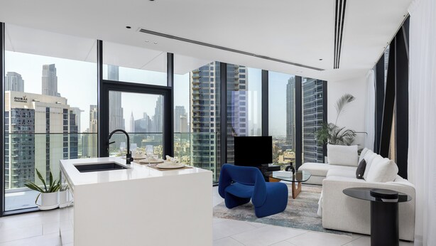 A modern living room with a white sofa, a blue chair, a small black table, and a kitchen area with a sink and a flat-screen TV. - Maison Privee - Sky-High Panoramic Apt with Iconic Burj Khalifa Views (Dubai)