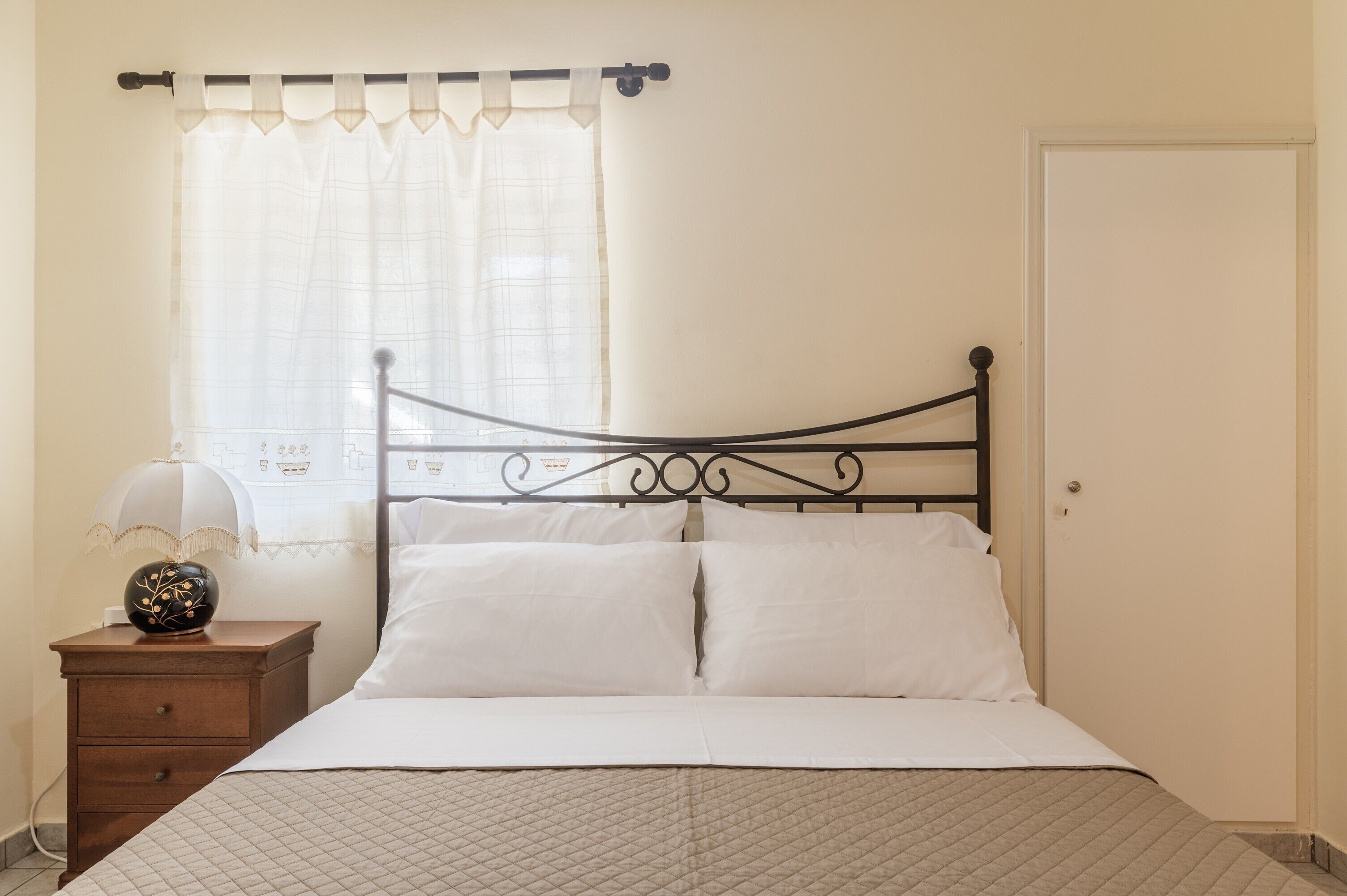 Ein Bett mit gesteppter Bettdecke, ein Nachttisch mit Lampe und ein Fenster mit durchsichtigem Vorhang.