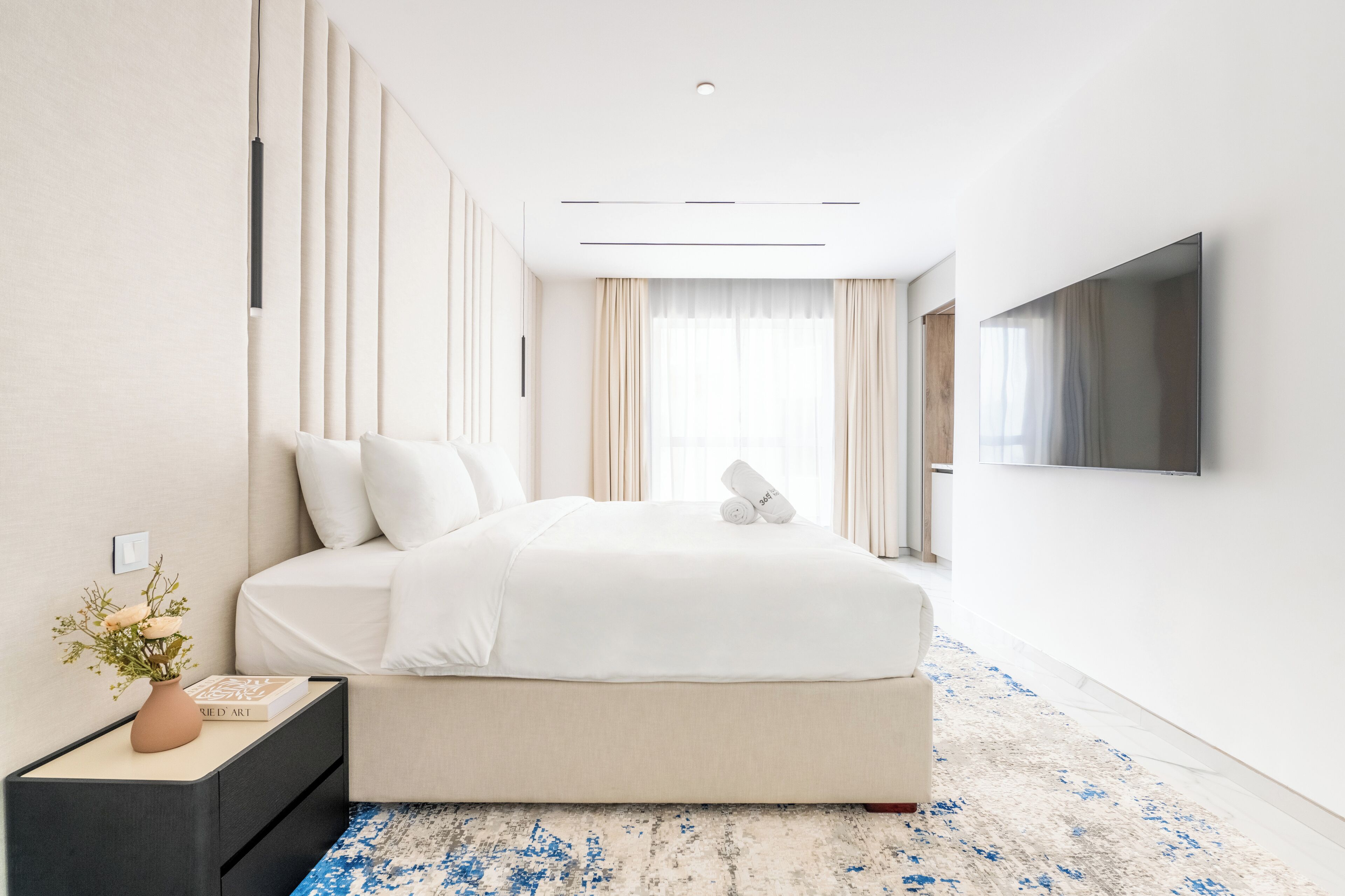 A modern hotel room with a large bed, a flat-screen TV mounted on the wall, a bedside table with a vase of flowers, and a large window with curtains.