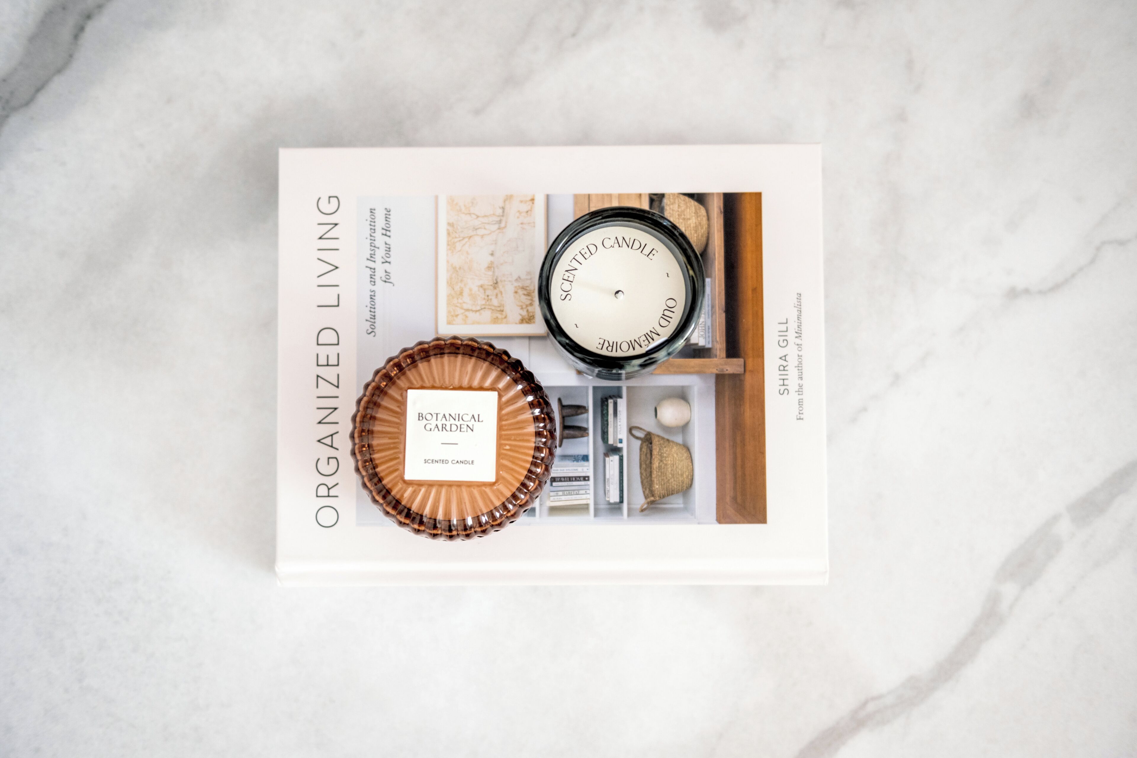 A book titled 'ORGANIZED LIVING' with a 'BOTANICAL GARDEN' scented candle and a 'SCENTED CANDLE' by Shira GILL on a marble surface.