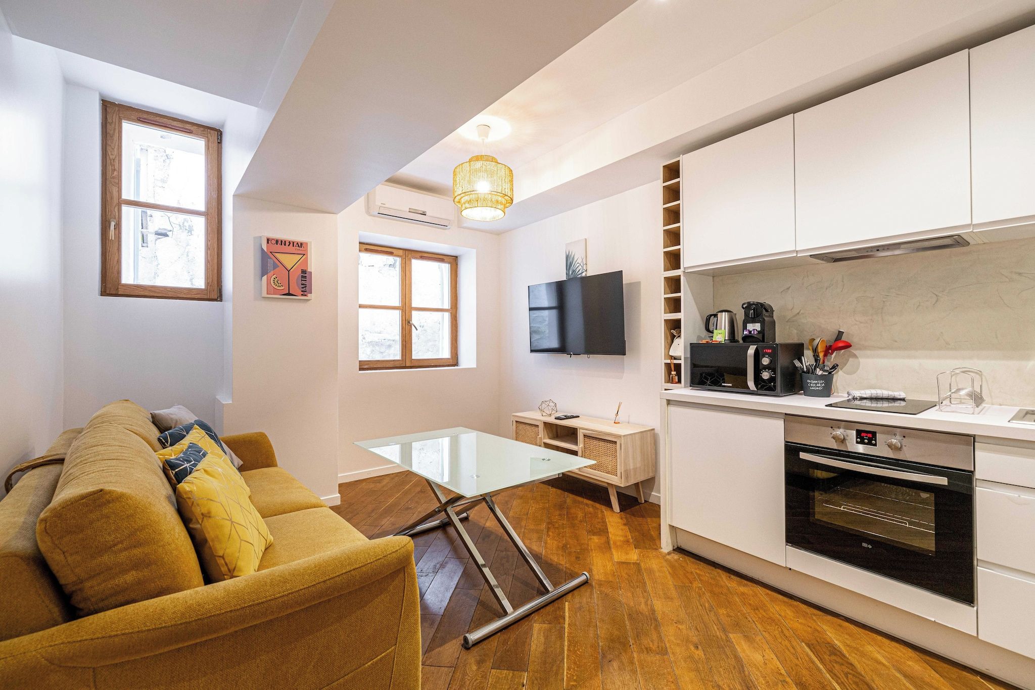 A compact living space with a sofa, a glass-top coffee table, a TV, a kitchen area with a stove, and a window.
