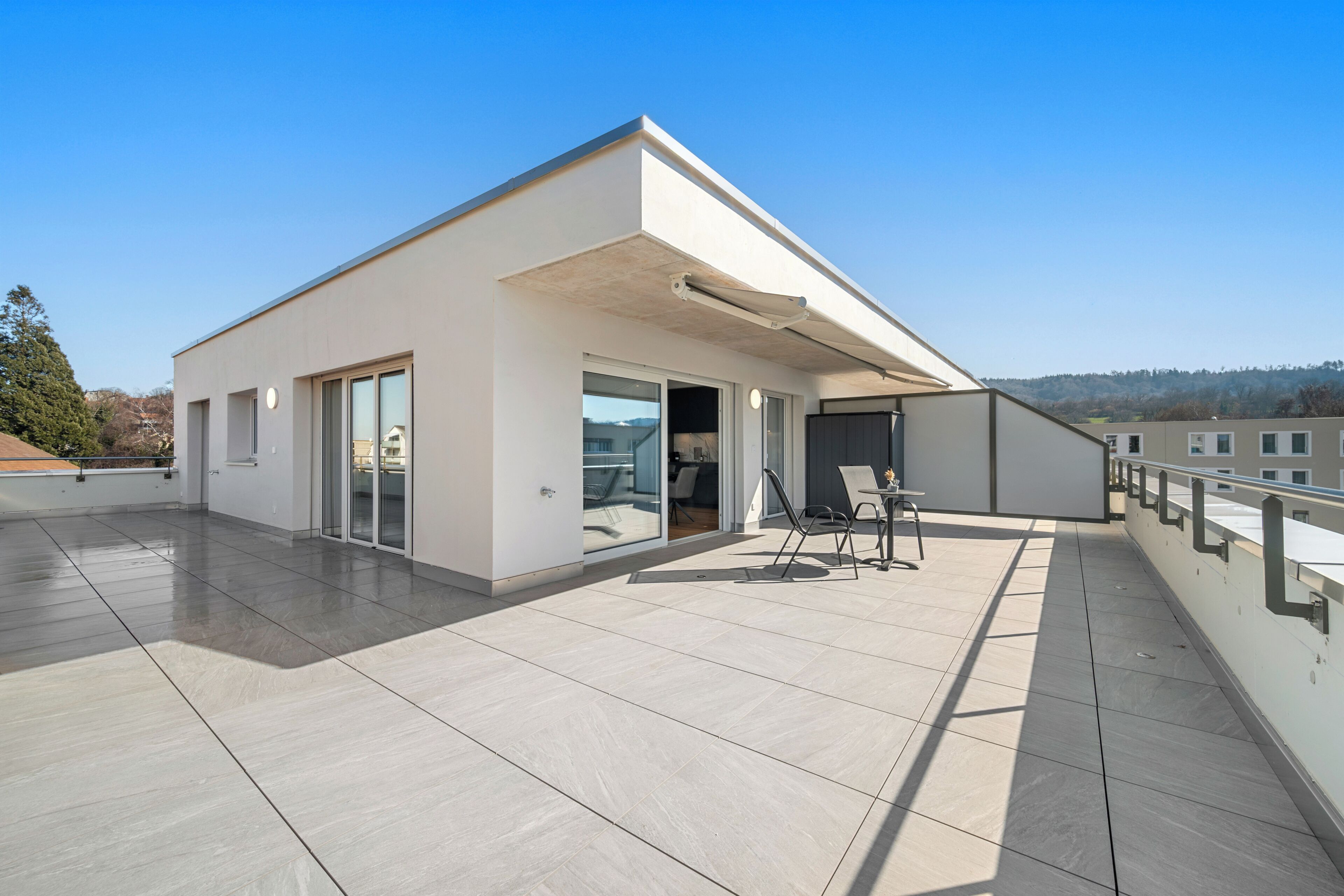 A modern building with a flat roof, large sliding glass doors, and a patio area with a table and chairs.