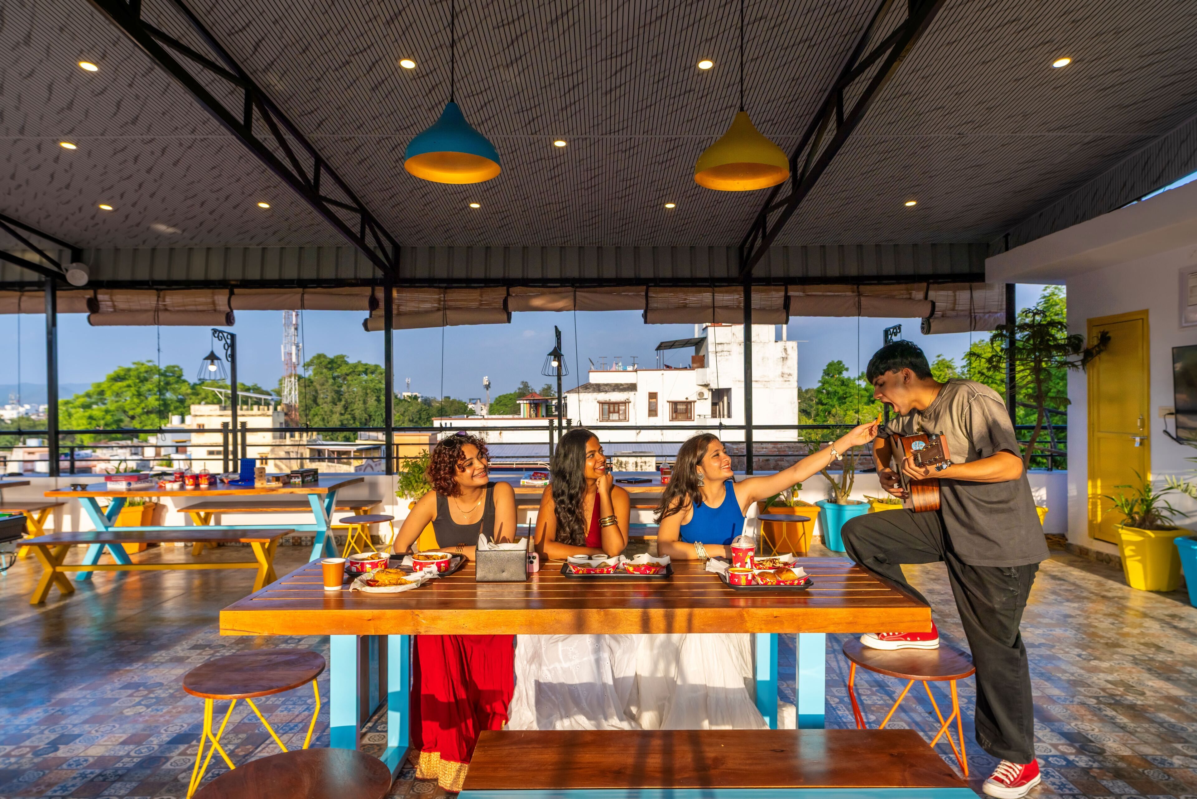 An outdoor dining area with wooden tables and benches, large windows, and hanging pendant lights.