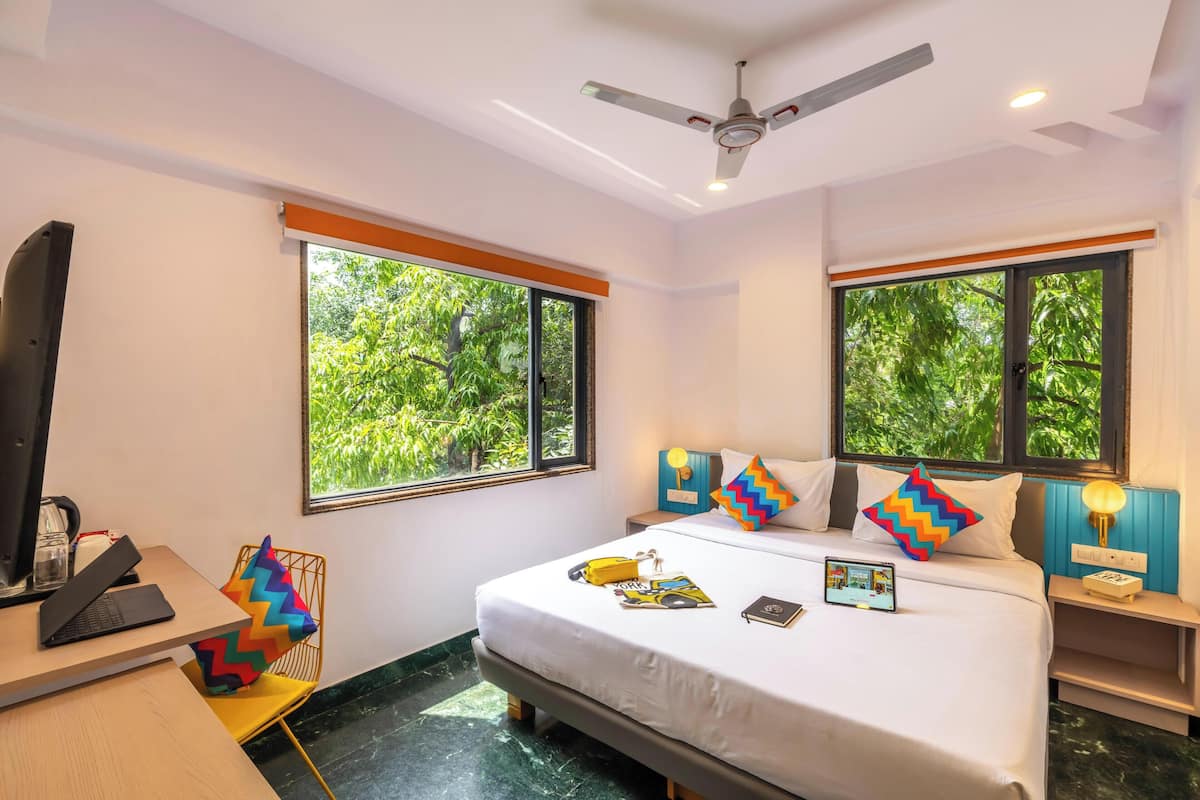 A hotel room with a bed, desk, chair, and large windows with a view of greenery.