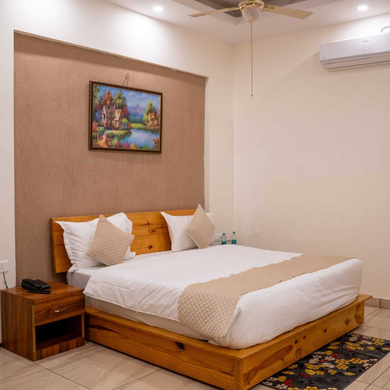 A bedroom with a wooden bed, white bedding, a nightstand, a painting on the wall, and an air conditioning unit.