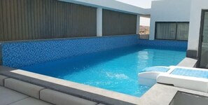 Ein Dachpool mit blauen Mosaikfliesen, eine kleine Lounge mit weißen Liegen und Blick auf die Stadt.