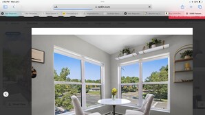 A room with a round table and two chairs, large windows with a view of a street and greenery, and a shelf with plants.