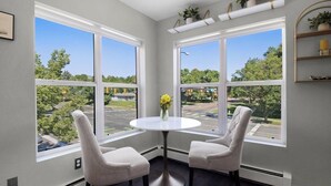 A corner room with two chairs, a small table, and large windows offering a view of a street and greenery. Shelves with plants are above the windows.