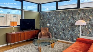 Un salon moderne avec une table basse en verre, un canapé en cuir marron, un téléviseur posé sur un meuble en bois et un papier peint à motifs.