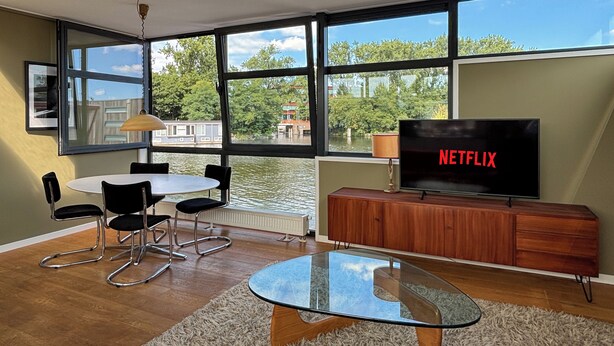 Un salon moderne avec une table basse en verre, un téléviseur posé sur un meuble en bois et de grandes fenêtres donnant sur un plan d’eau.