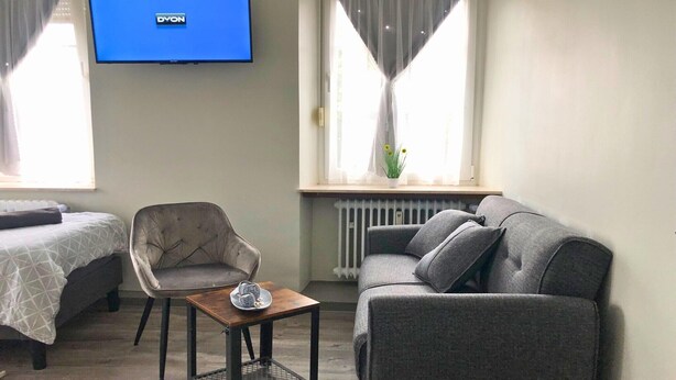 A room with a bed, a grey armchair, a grey sofa, a small table, and a TV mounted on the wall. - Apartment in Pellingen near Saar-Hunsrück (Pellingen)
