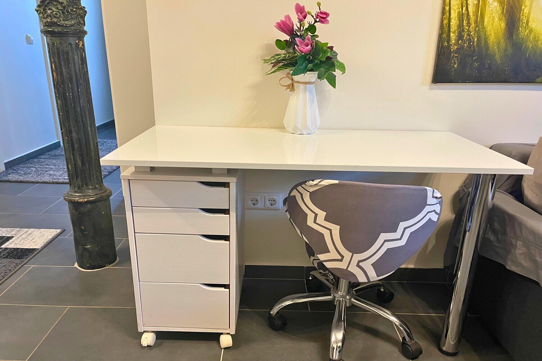 A white desk with a vase of flowers, a modern office chair, and a white cabinet with drawers.
