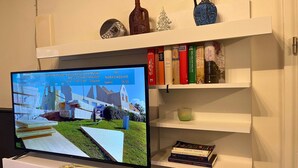 A television displaying a news channel with a cityscape background, a white shelf with decorative items and books, and a white cabinet below the TV.