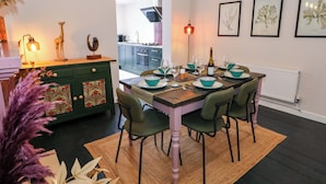 Un coin repas avec une table en bois dressée pour six personnes, entourée de chaises vertes, et une cuisine en arrière-plan.
