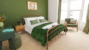 Une chambre bien rangée avec un mur vert, un lit recouvert d’une couverture verte, une chaise, un bureau avec un livre et une fenêtre avec des rideaux.