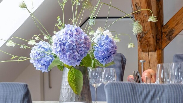 A dining table with a vase of flowers, a hanging light fixture, and a wooden ceiling beam. - Ahoy Amore (Sylt)