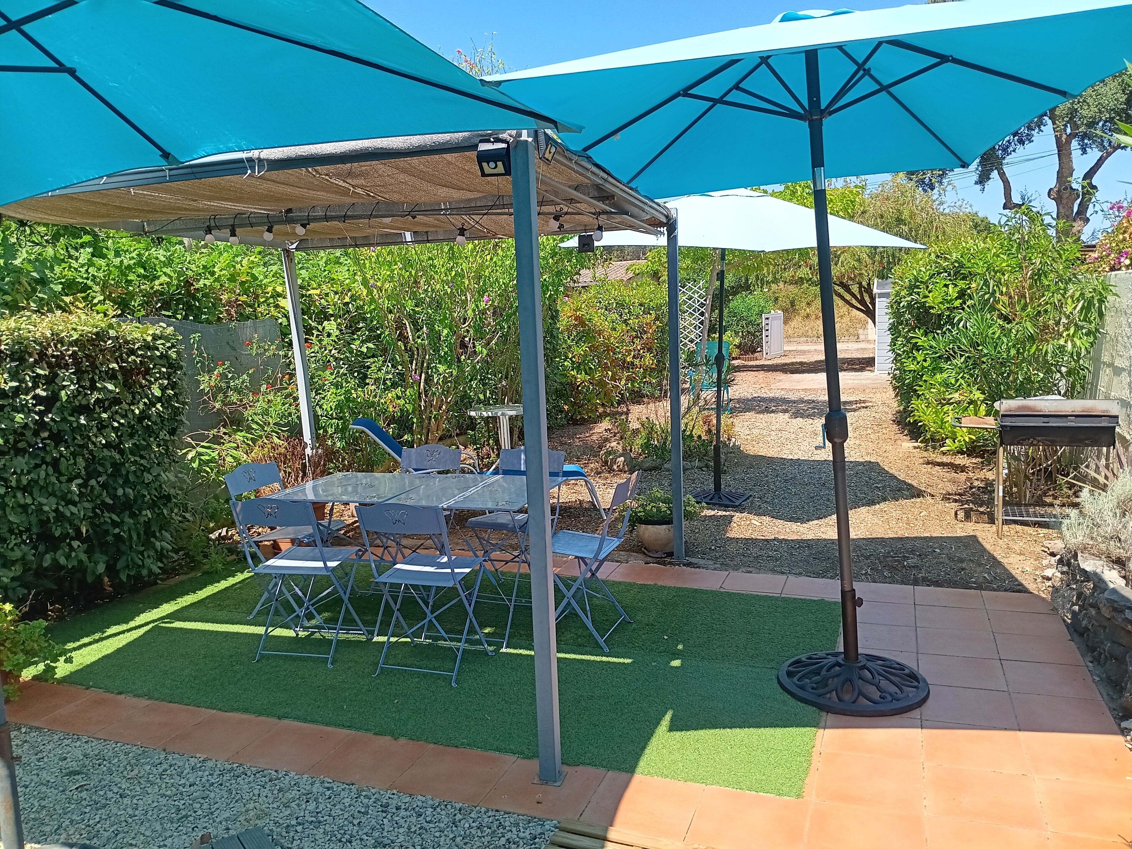 A patio area with a table and chairs under a blue canopy, surrounded by greenery and a gravel path.
