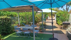 A patio area with a table and chairs under a blue canopy, surrounded by greenery and a gravel path. - Corse, San giuliano, 50 kms de Bastia, maison à 2mn de la mer, animaux acceptés (san giuliano)