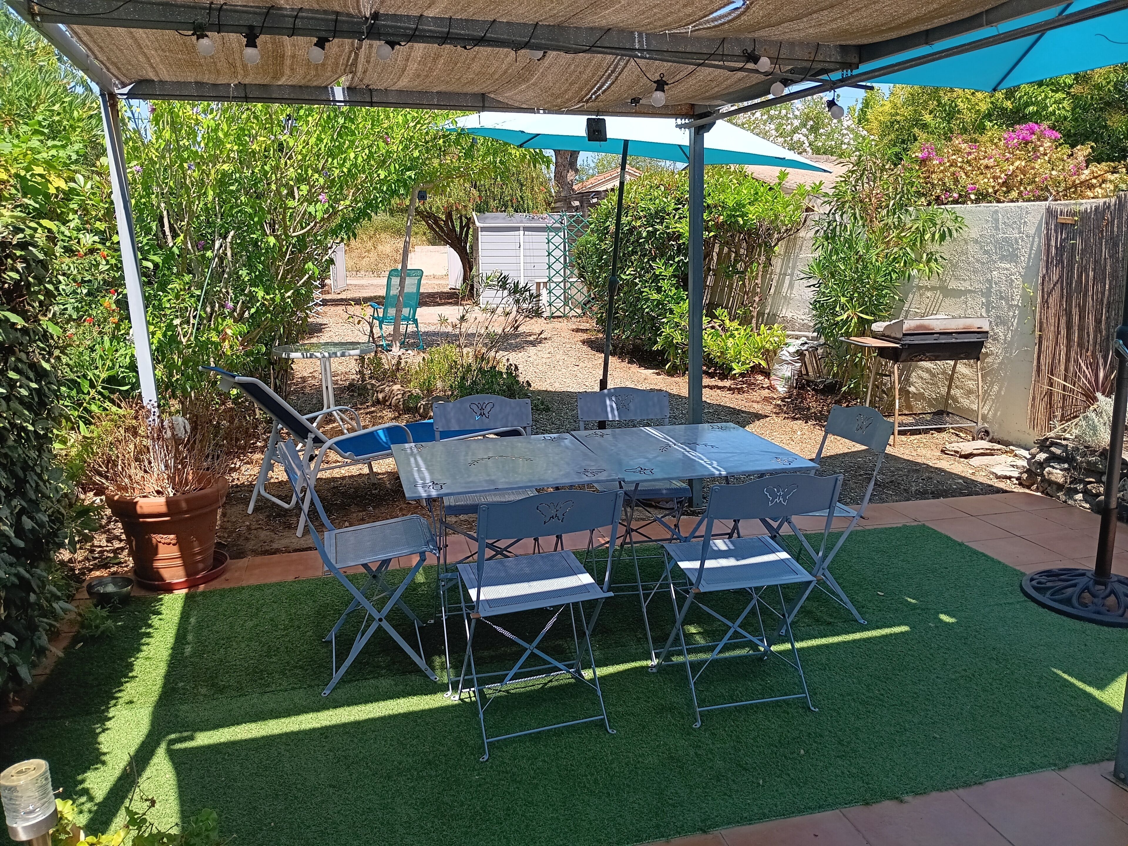 En overdækket terrasse med et bord, stole, grøn bevoksning og en grill.