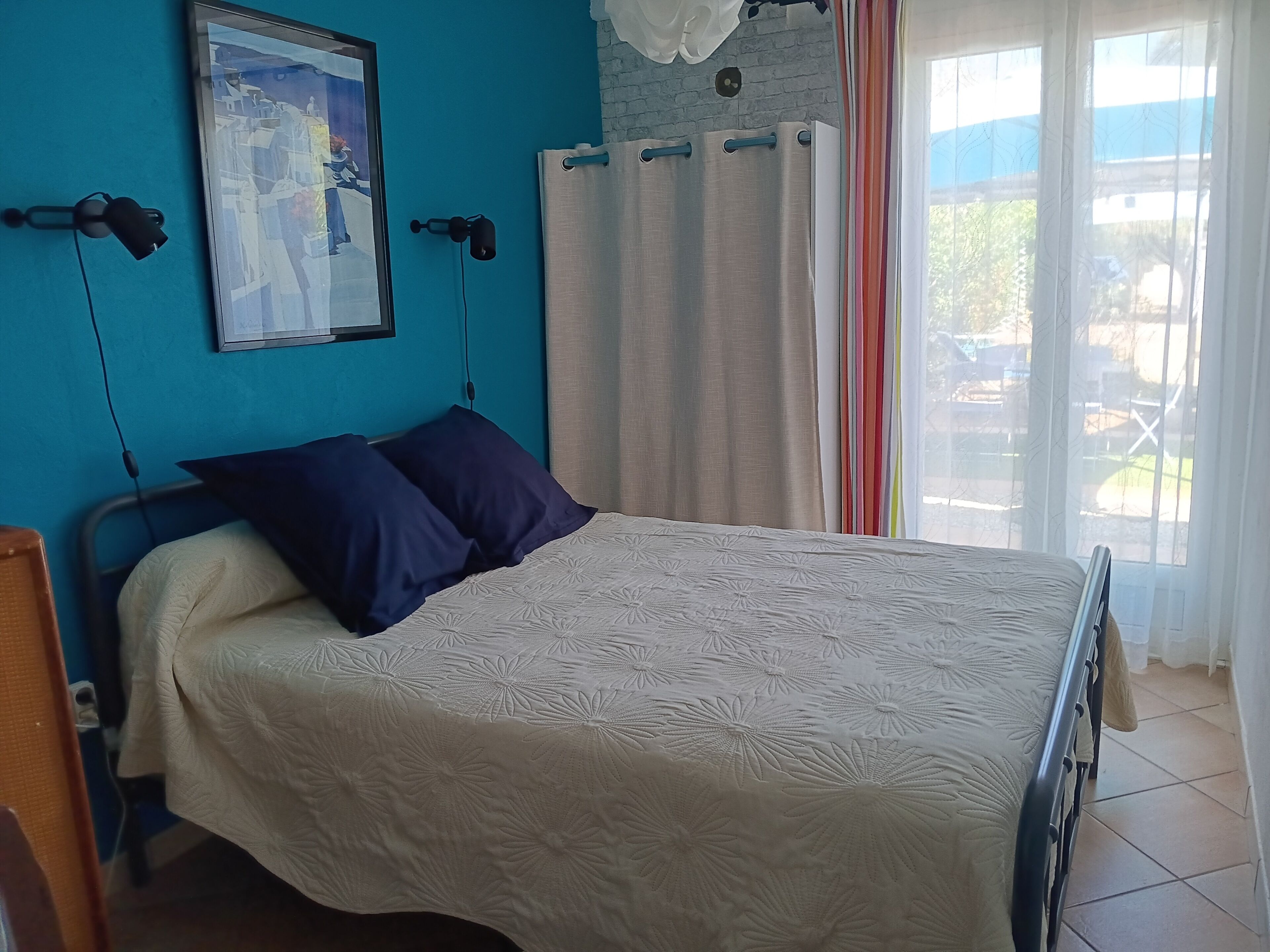 A bedroom with a bed, a blue wall, a window with curtains, and a framed picture on the wall.