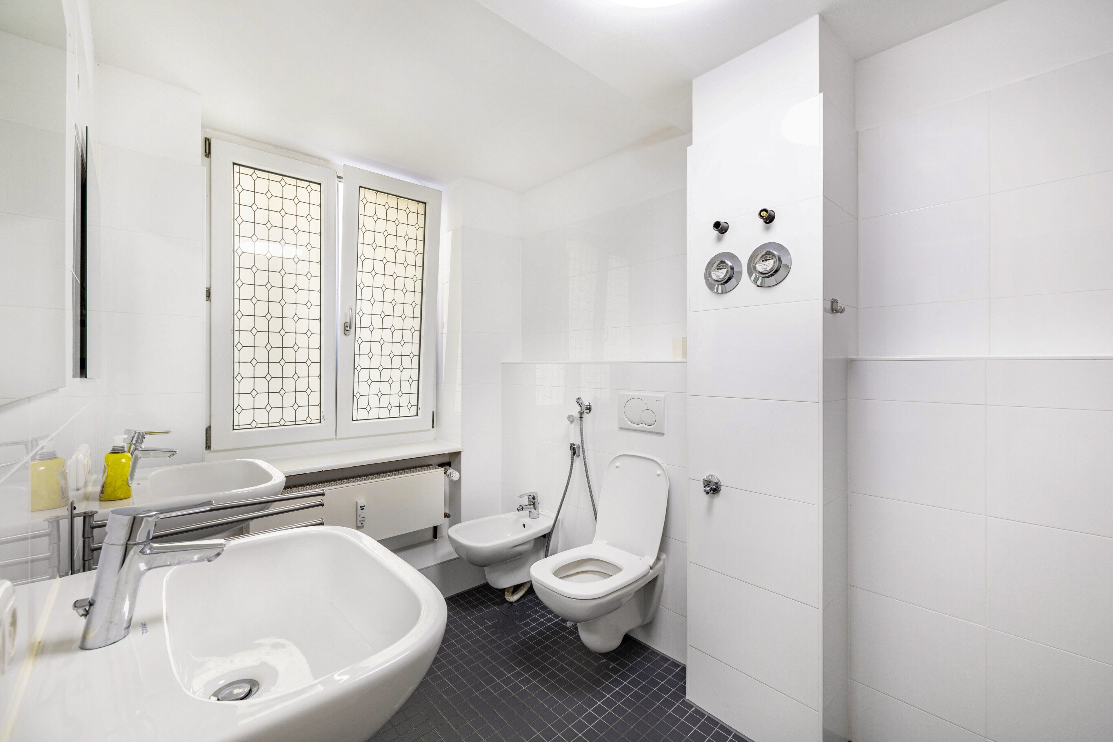 Une salle de bain moderne équipée d’un lavabo, d’un bidet, de toilettes et d’une fenêtre ornée d’un motif géométrique.